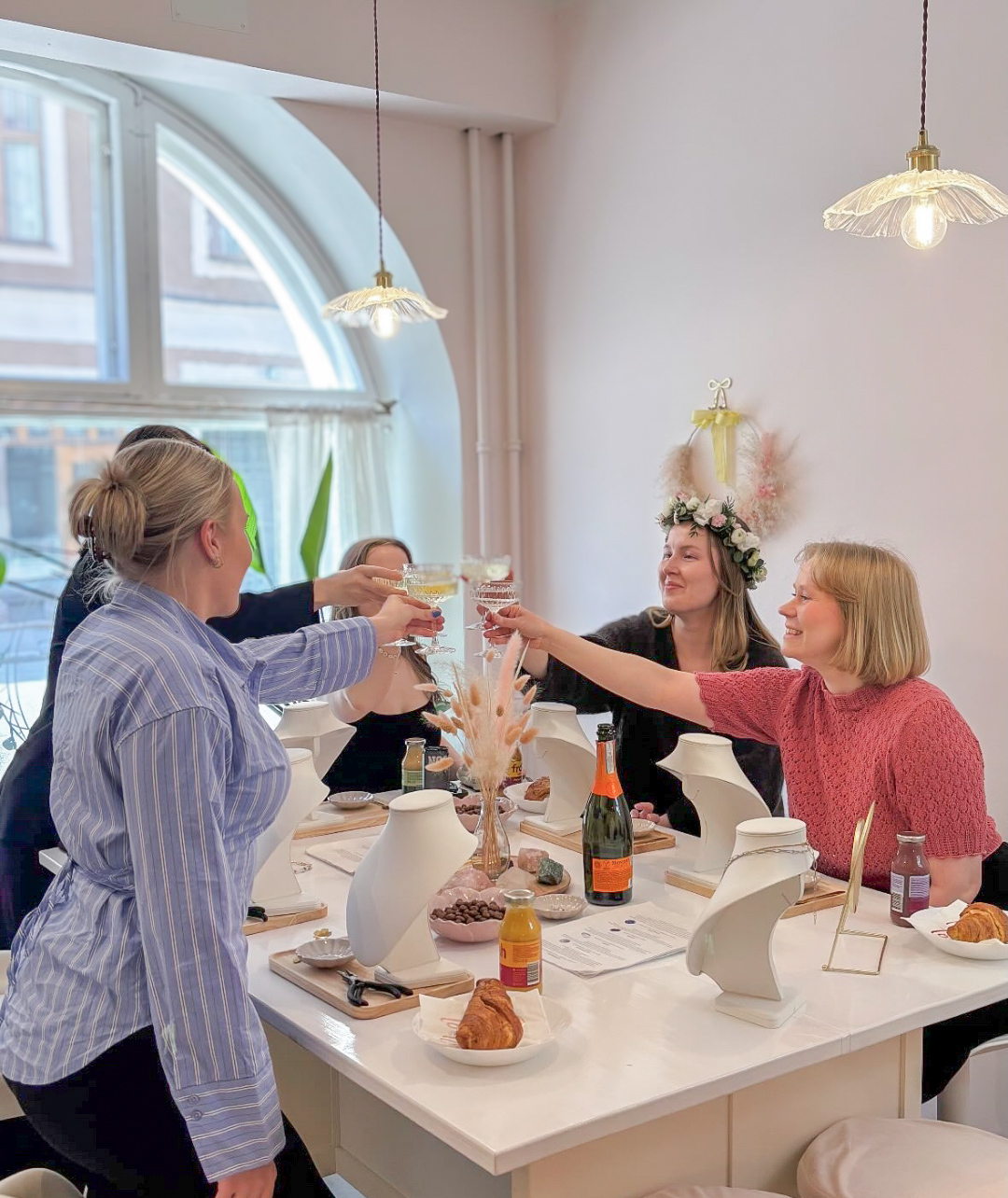 Naiset juhlapöydässä nostavat laseja, koristeelliset kattokruunut ja ikkunakaari taustalla. Yhteinen hetki Portal of Joy:n pastellisen sävyisessä myymälässä Helsingissä.