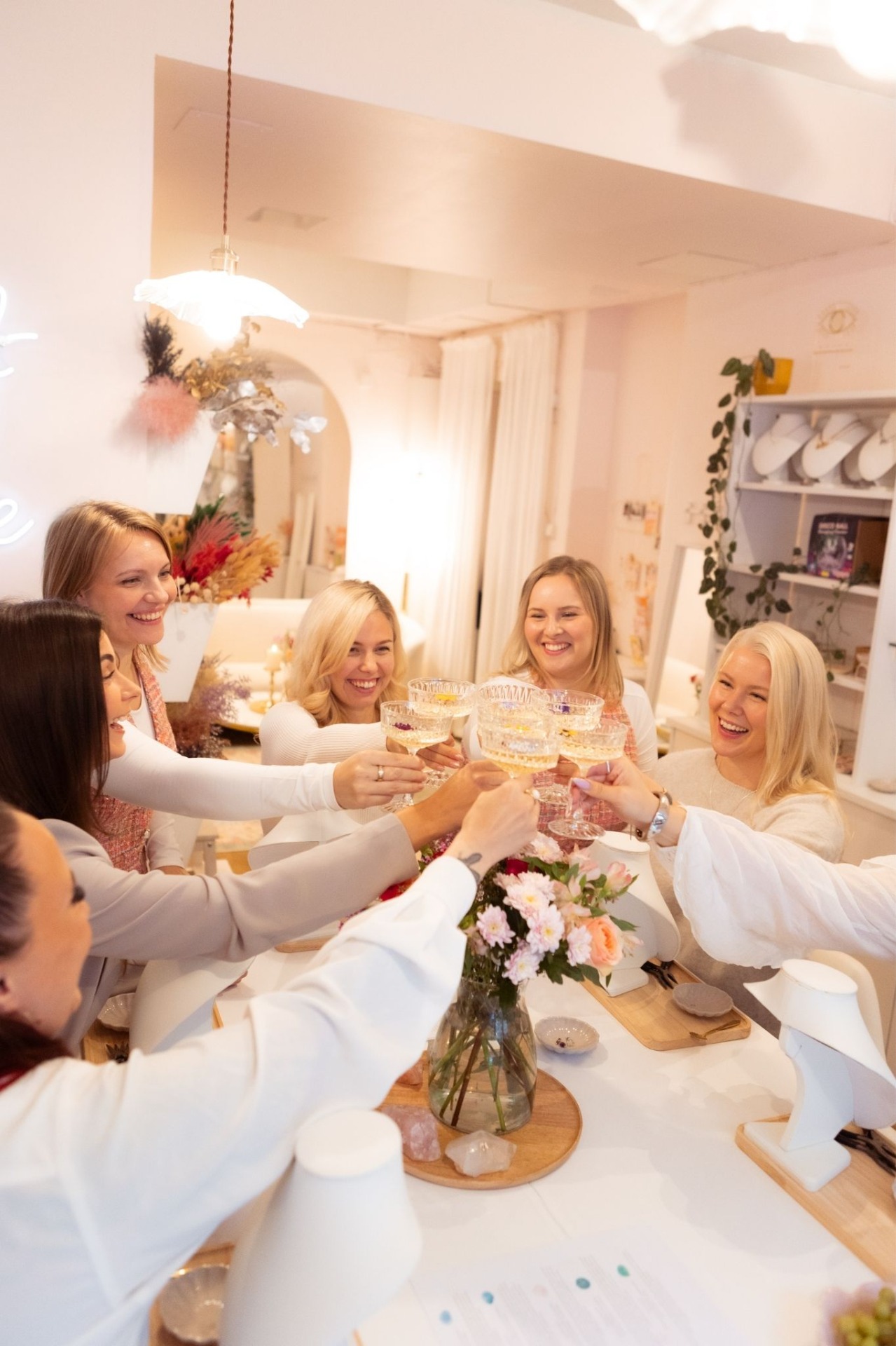 Naiset juhlapöydässä nostavat laseja, koristeelliset kattokruunut ja ikkunakaari taustalla. Yhteinen hetki Portal of Joy:n pastellisen sävyisessä myymälässä Helsingissä.